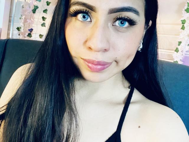 Close-up of ZoeRedds, a woman with long black hair, blue eyes, and full pink lips, wearing a black top. She's looking directly at the camera with a slight smile, creating a sexy vibe.