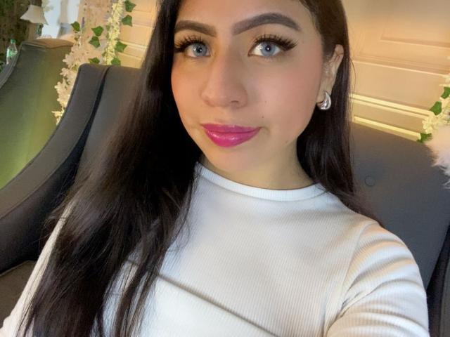 ZoeRedds has long black hair, blue eyes, and pink lipstick. She's wearing a white ribbed top, sitting in a black chair. Her makeup is heavy, with thick black eyeliner. She's looking directly at the camera with a slight smile.
