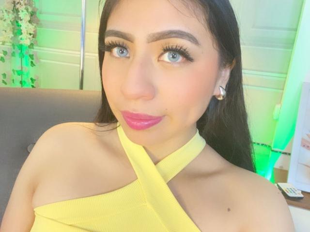Close-up photo of ZoeRedds, a sexy woman with long black hair, blue eyes, and pink lipstick, wearing a yellow halter top. She has a sultry expression, green light in background.