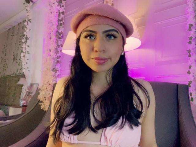ZoeRedds with long black hair, pink beret, and white top, sitting in a chair with purple lighting. She's giving a slight smile, looking directly at the camera, with visible sexual tension.