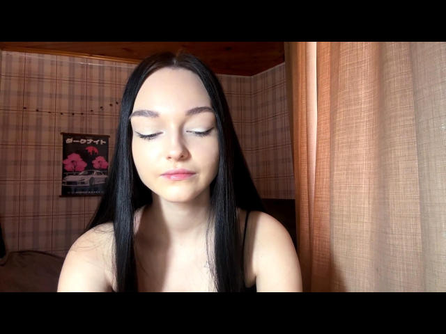 Digital photo of RoseanaConlin, fair-skinned woman with long black hair, closed eyes, and pink lips, sitting in a room with wooden walls and curtains. Her chest is exposed, showing cleavage.