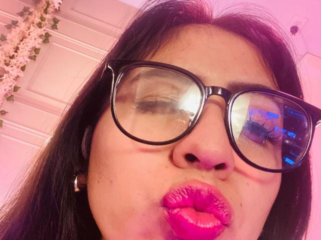 Nahaufnahme-Selfie von ZoeRedds mit dickem, schwarzem Haar, großen, schwarzen Brillen und leuchtend rosa Lippenstift. Sie zieht die Lippen zusammen und reflektiert rosa und violettes Licht. Ihr Gesicht ist im Mittelpunkt, der Hintergrund ist unscharf..