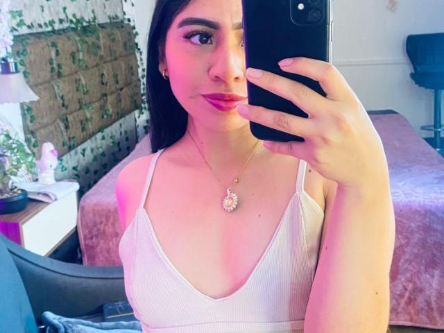 ZoeRedds, maakt een foto van zichzelf in de spiegel met een zwarte telefoon, draagt een witte ribtrui en laat veel decolleté zien, heeft donker haar. Rode lippenstift en een gouden ketting. Achtergrond voor een slaapkamer met een roze bed..