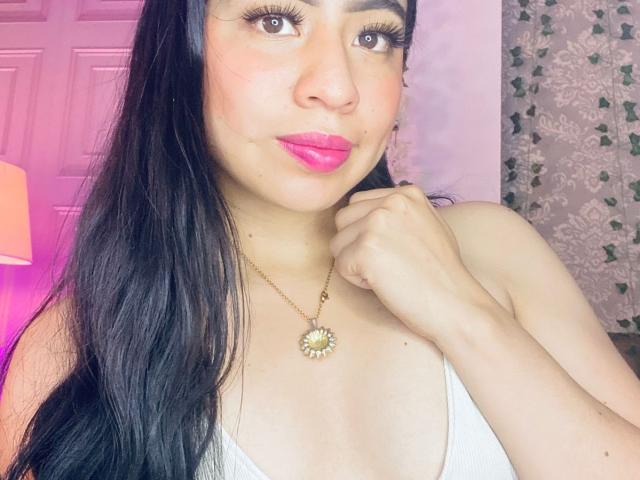 ZoeRedds has long black hair, pink lipstick, and a sun pendant necklace. She's touching her neck, with a white top. Bright lighting, pink and purple hues. Sexual tension, seductive gaze.
