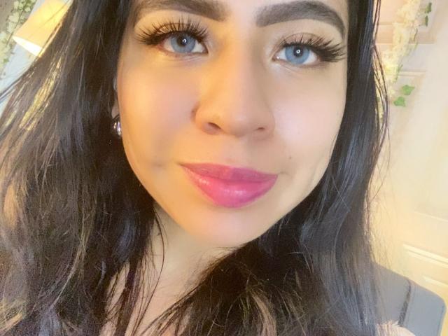 Close-up de ZoeRedds, uma jovem com cabelos longos e pretos, olhos azuis e batom rosa. Ela tem sobrancelhas grossas, cílios longos e está sorrindo levemente. Seu olhar é direto e sedutor..