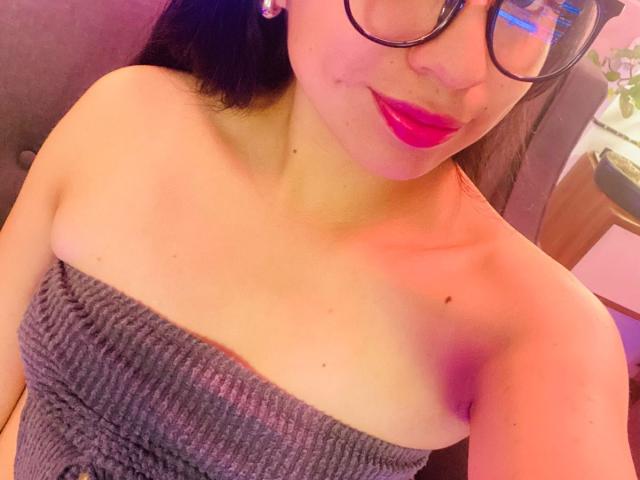 Close-up of ZoeRedds, an Asian woman with black hair, wearing glasses and a strapless, textured, gray top. Her red lips are slightly parted, showing a hint of a smirk. Her breasts are exposed.