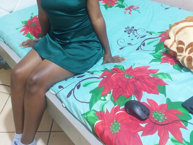 WandaCandy está sentada en una cama con sábanas florales de color azul brillante, vestida con un vestido verde ajustado que deja ver sus muslos morenos. Un control remoto negro está sobre la cama, cerca de su lado derecho..