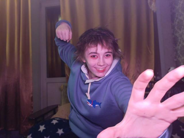 KarisaWinkel, met rommelig bruin haar, met een grijns, gekleed in een blauwe hoodie, reikt met een gloeiende hand uit, met een opgetrokken vuist. Een zwak verlichte kamer met bruine gordijnen, sterren op de vloer. Heel speels..