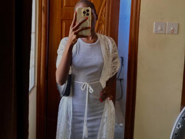 KharlaQueen, a woman with tan skin, stands in front of a mirror, holding a phone in front of her face. She's wearing a tight grey dress and a white lace robe, exposing her medium-sized breasts. Her right hand grips her robe, showing off a visible nipple.