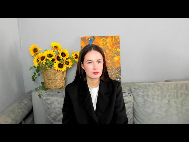 Hizer, with long dark hair and pale skin, sits on a gray couch, wearing a black blazer over a white shirt. Sunflowers in a basket and an orange painting in the background.