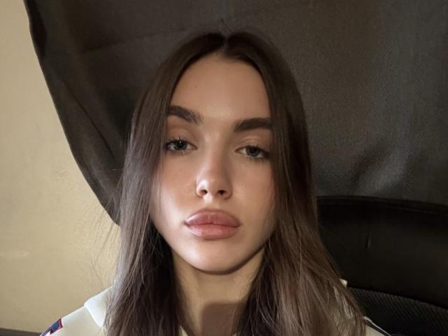 Gros plan d'EmeraldWeinreb, une femme aux longs cheveux bruns, aux lèvres pulpeuses et aux sourcils foncés, qui regarde directement l'appareil photo. Elle porte un haut blanc, et des ombres mettent en valeur son visage. Extrêmement sexy..