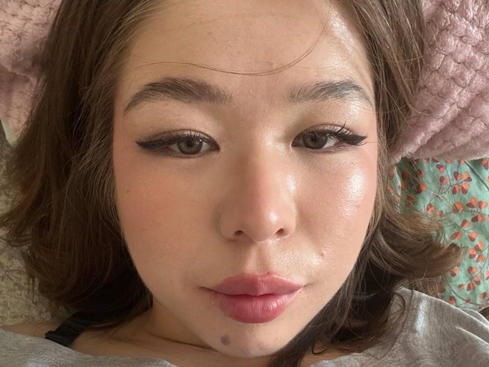 Primo piano di BelkisMuldoon, il cui viso è illuminato dalla luce del sole, con capelli castani, eyeliner nero, un leggero sorriso e una piccola lentiggine sotto il labbro. pelle leggermente lucida, indossa una camicia grigia, cuscino rosa chiaro sullo sfondo..