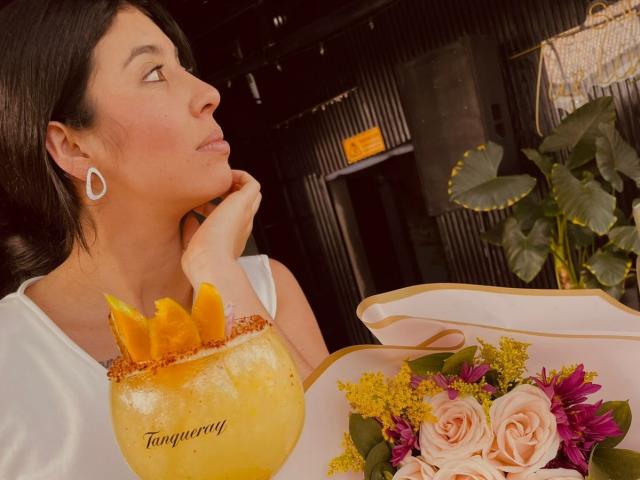 MiluSky, con cabello oscuro y vestido blanco, mira hacia un lado mientras apoya su barbilla en su mano. Ella sostiene un tazón amarillo con rodajas de naranja y un arreglo floral rosa y blanco..