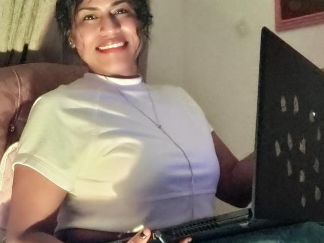 Foto van IvannaDuran zittend op een bruine stoel, met een glimlach en witte tanden, gekleed in een strak wit T-shirt. benadrukt haar grote borsten. Laptop voorin, zonlicht werpt schaduwen.