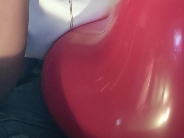 Close-up of a glossy red latex balloon pressed against IvannaDuran's buttocks, creating a shiny reflection. The balloon's tight fit suggests a sensual, erotic context. Bright light in the background.