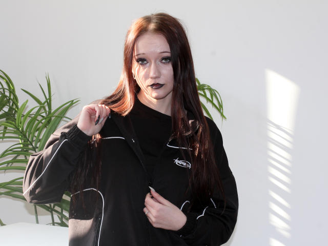 OliviaGilson stands in bright light, wearing a black hoodie with white stripes, holding her hair with one hand and gripping her chest with the other. Her dark lipstick contrasts with her pale skin.