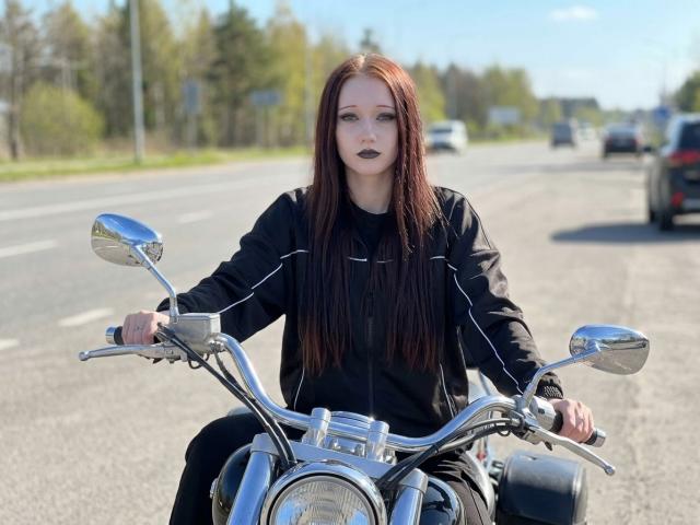 Olivia Gilson, avec ses longs cheveux roux et son rouge à lèvres noir, est assise sur une moto chromée, agrippant le guidon. Elle porte une veste noire, et on peut voir une autoroute et des arbres en arrière-plan. Extrêmement sexy..