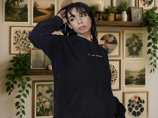 KatherinJohns, une femme aux formes généreuses avec des cheveux noirs, se tient devant un mur orné de cadres d'œuvres d'art, vêtue d'un sweat à capuche noir, une main cheveux, regard séduisant, œuvre d'art encadrée, plantes, bougies.