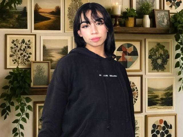 Katherin Johns está em frente a uma parede coberta com impressões botânicas emolduradas e plantas em vasos. Ela tem cabelo preto, veste um moletom preto e olha diretamente para a câmera. Muito quente..