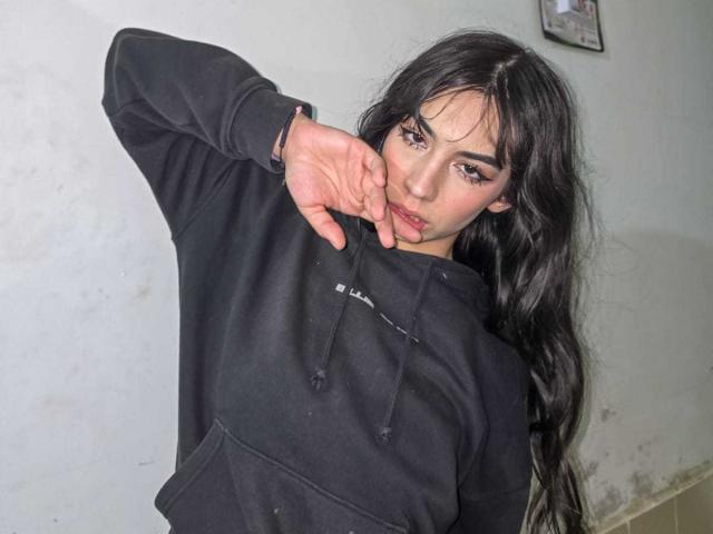 Photograph of KatherinJohns, wearing a black hoodie, with long black hair, dark eyebrows, and red nails, sucking on her fingers, leaning forward with a sultry expression, white wall background.