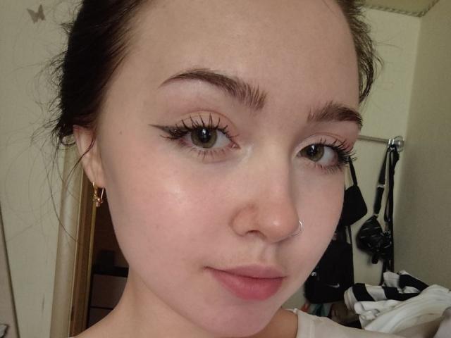 Close-up selfie of TerraMaul, a young woman with fair skin, dark eyebrows, and long eyelashes. She has a nose ring, small gold hoop earrings, and subtle pink lips. Her expression is neutral.