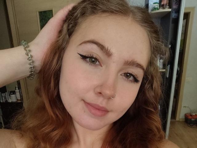 Close-up de TerraMaul, com pele clara, cabelo castanho ondulado, delineador preto e um piercing no nariz. Ela está sorrindo, com a mão direita no cabelo, e usando uma pulseira de prata. Muito atraente..