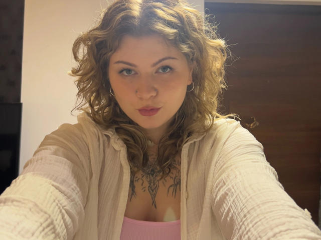 AlinaHeiss-hot, with wavy brown hair and red lipstick, stares directly at the camera, wearing a white cardigan over a pink top, revealing a large chest tattoo, in a brightly lit room.