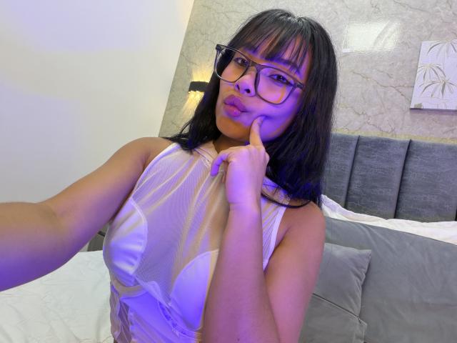 Fotografía de Vannesa Saenz con cabello negro hasta los hombros y gafas, vistiendo una blusa blanca de encaje, tocándose la barbilla con un dedo. Está en una cama gris con almohadas blancas, luciendo seductora..