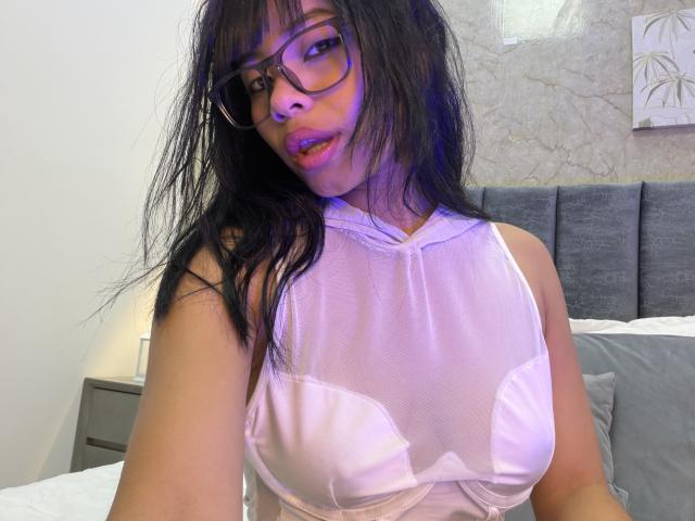 VannesaSaenz, une femme latina aux longs cheveux noirs et portant des lunettes, porte un haut blanc transparent qui met en valeur sa poitrine généreuse. Elle est assise sur un lit, dégageant une tension sexuelle, et regarde l'appareil photo..