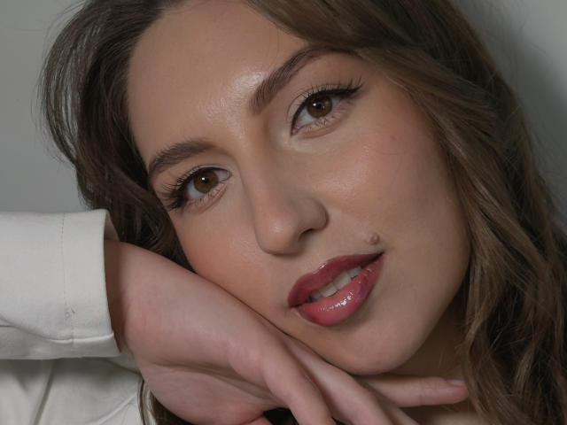 Primer plano de LoriSue, con el rostro ligeramente inclinado, ojos marrones que miran seductoramente a la cámara, labios rosados y brillantes entreabiertos, a la derecha. Mano con los dedos extendidos sobre su mejilla, cabello ondulado castaño, camisa blanca..