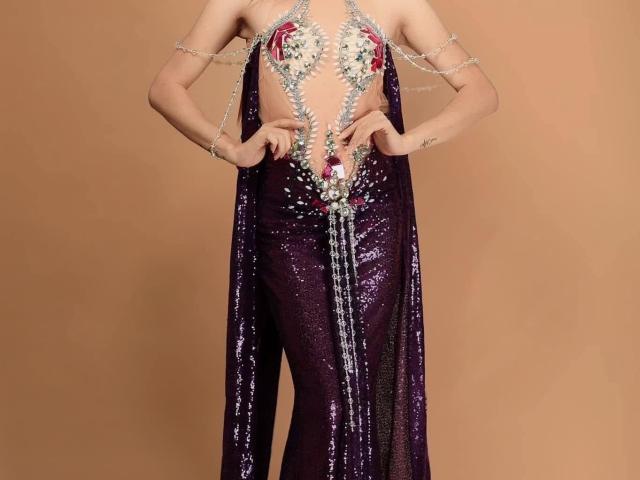 AdrianaLuxe vestindo um macacão roxo brilhante com decote baixo e um profundo decote em V adornado com correntes de prata, segurando o correntes na frente, revelando lingerie vermelha, fundo marrom. Pose sensual e provocante.