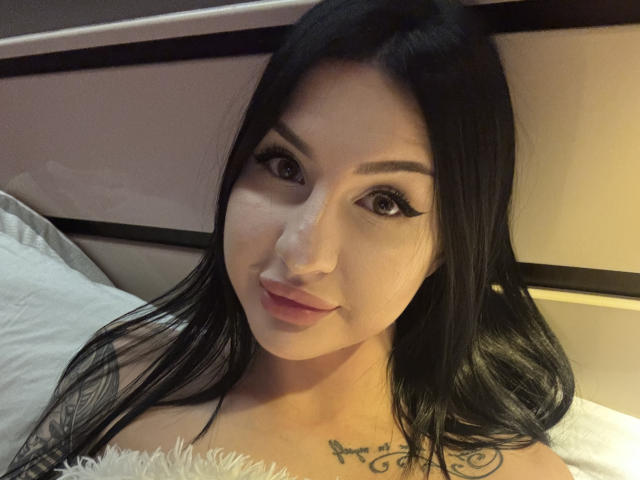 Gros plan d'AuraXo, une femme asiatique aux longs cheveux noirs, avec un trait d'eye-liner prononcé et des lèvres pulpeuses, allongée sur un lit blanc. coussin, tatouage visible sur la poitrine, lumière douce, tension sexuelle.