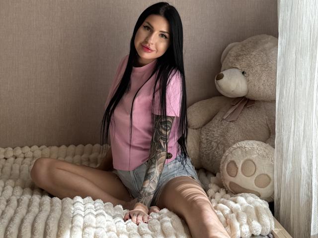 AuraXo, una donna dai capelli scuri e lunghi neri, è seduta su una coperta bianca con una trama particolare, indossando una camicia rosa e jeans. pantaloncini. Grande orsacchiotto beige a destra. Estremamente attraente, posa rilassata..