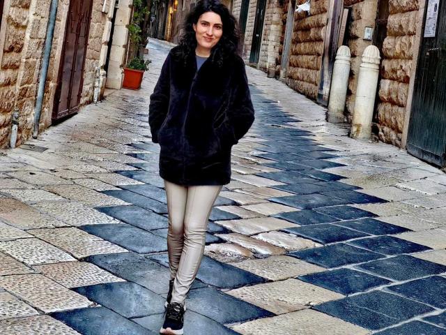 A AddictedToUTS caminha por um beco de pedra quadriculado, vestindo um casaco de pele preta, leggings brancas e sapatos pretos. Ela tem cabelos cacheados escuros e um sorriso confiante. O asfalto molhado reflete a luz..