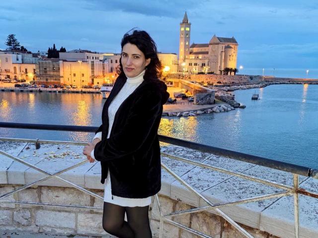 AddictedToUTS stands in a snowy coastal town, wearing a black coat over a white dress, black stockings, and black gloves. Historic buildings and a lit tower in the background. Fucking sexy.