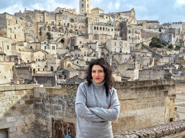 AddictedToUTS with dark curly hair, red lipstick, and crossed arms stands in front of a bustling, sunlit stone village. Stone buildings fill the background. Bright, clear day. Casual, confident stance.