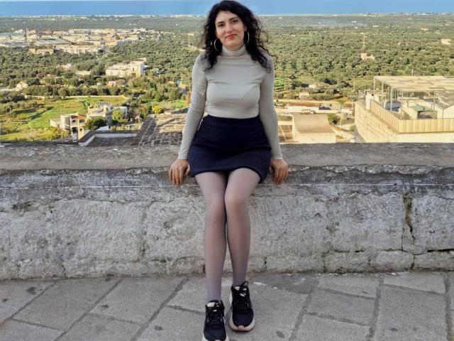 AddictedToUTS, uma mulher curvilínea com pele clara e cabelos cacheados escuros, está sentada em um degrau de pedra, vestindo uma gola alta branca. Saia preta e tênis pretos, com um fundo que mostra uma paisagem urbana. Muito atraente..