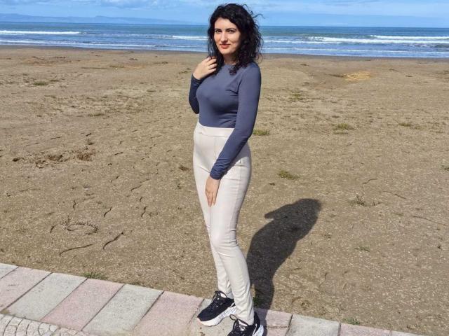 AddictedToUTS se tient sur une plage, portant un haut bleu marine ajusté à manches longues et un pantalon blanc, ainsi que des baskets noires. Elle a les cheveux noirs et bouclés, une expression confiante et une ombre visible. Fond d'écran avec un océan.