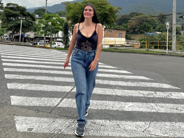 LizzaRose, de pie con confianza en un paso de cebra, vestía una camiseta de encaje negra y pantalones vaqueros azules, con zapatillas grises. Su cabello oscuro fluye libremente. Fondo: árboles, edificios, montañas. Tensión sexual: atracción visible..