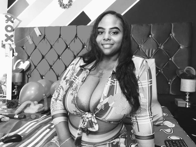 Zwart-wit foto van Hayley Evans, volslank met grote borsten, die een opgetrokken shirt draagt en op een bed met een stoffen bekleding zit. hoofdeinde. Ballonnen en een lamp op de achtergrond. Sensuele, zelfverzekerde pose..