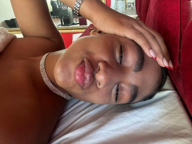 Close-up photo of NatalyBlonder with her head resting on a white pillow, hand on forehead, wearing a silver choker and bracelet, lips glossy, eyes half-open, red nails, sexy, relaxed expression.