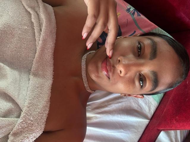 NatalyBlonder, with tan skin and dark hair, lies on a bed, wearing a white towel wrapped around her chest. She has a seductive expression, touching her lips with one hand.