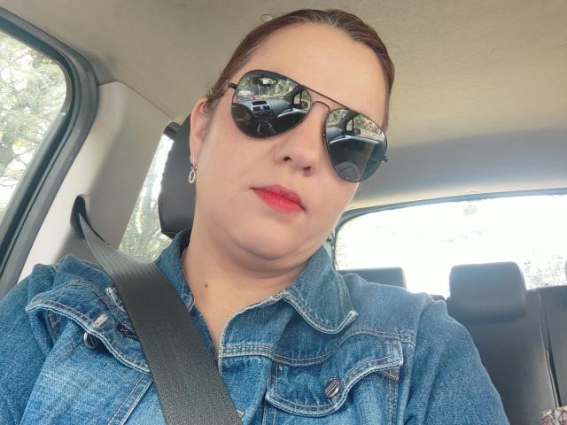 Amelia Wilson, met bleke huid en rode lippenstift, die zwarte zonnebril draagt en een blauw denim shirt, zittend in een auto met een de veiligheidsgordel over haar borst, weerspiegeld in de spiegel..