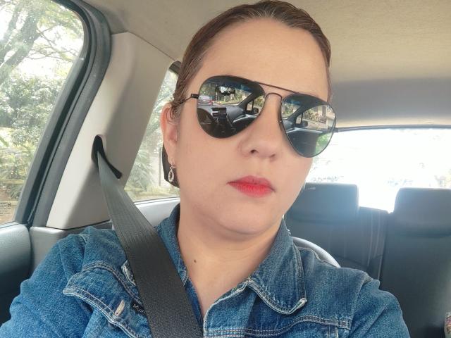 Close-up van Amelia Wilson, zittend in een auto, met reflecterende zonnebril, rode lippenstift en een blauw denim shirt. De veiligheidsgordel zat over haar borst. Haar uitdrukking is serieus en geconcentreerd..