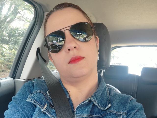 Amelia Wilson zit in een auto, ze draagt een blauw denim shirt en een zwarte zonnebril, met een veiligheidsgordel om haar borst, en rode lippenstift. lichte huid, reflectie in de bril, helder daglicht buiten, serieuze uitdrukking.