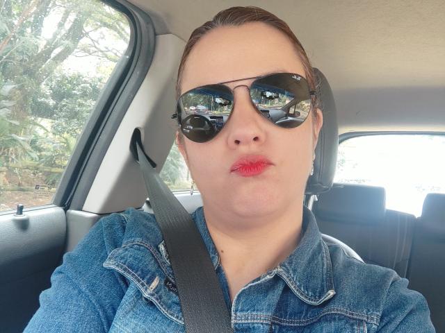 Amelia Wilson in de auto, draagt een zwarte zonnebril, rode lipstick en een blauw denim shirt, met een veiligheidsgordel om haar borst, en er komt licht binnen. Van links naar rechts: serieuze uitdrukking, lichte seksuele spanning, zichtbare reflectie in de zonnebril..