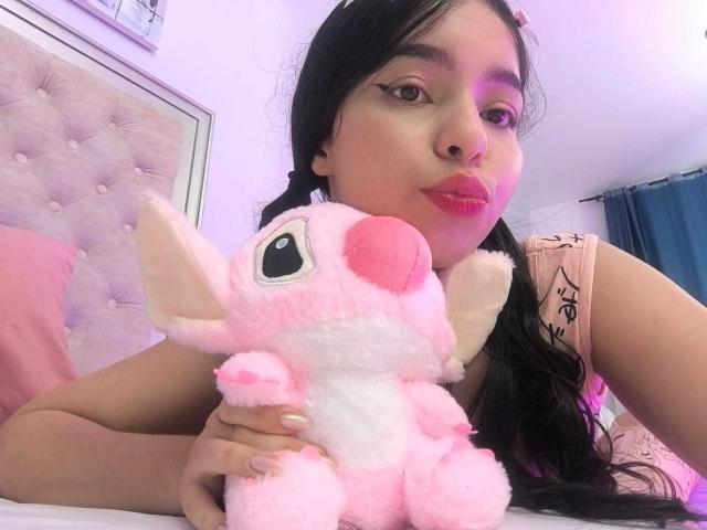 Lissy Carter, una giovane donna con lunghi capelli neri e rossetto rosso, tiene in mano un soffice giocattolo rosa con un grande occhio. Si sporge in avanti, indossando una canottiera rosa. Tensione sessuale.