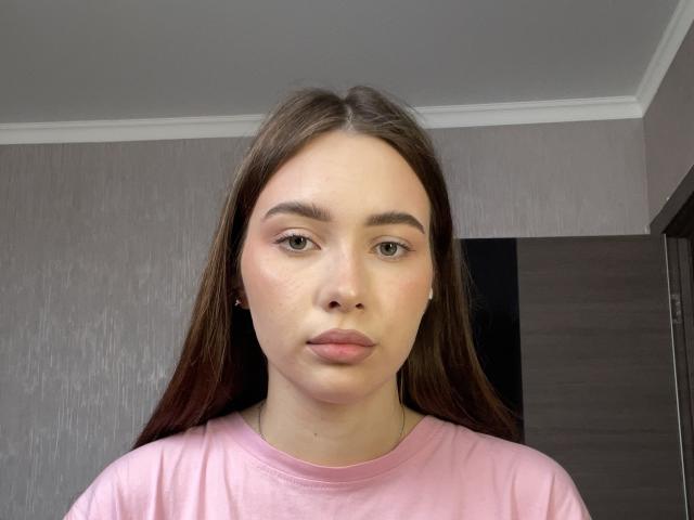 Photographie de Kenisha Haislett, avec de longs cheveux bruns foncés, portant une chemise rose, regardant directement l'appareil photo. Expression neutre.
lumière naturelle douce, fond texturé gris, ombre subtile sur la droite..
