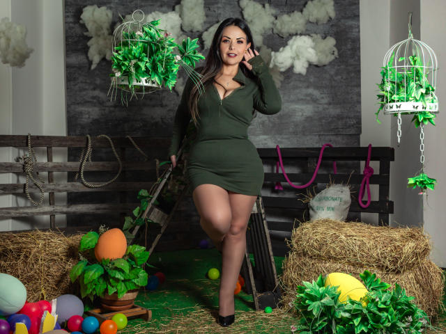 MelissaLeen, sexy, aux formes généreuses, robe verte foncée, manches longues, debout, main sur la hanche, bottes de foin, ballons colorés, suspendus. plantes, clôture en bois noir, nuages blancs, feuillage vert, regard intense, pose séduisante.