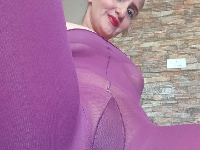 Close-up shot of RubiAnderson wearing a sheer purple bodysuit, showing her crotch with visible fabric lines. RubiAnderson has red lipstick, smiling down at the camera. Background: textured brick wall.